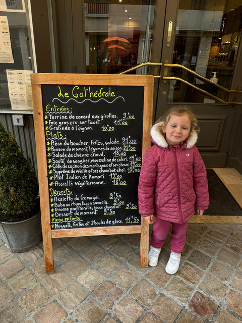 Jeune fille souriante devant le tableau de menu extérieur de Le Cathédrale Brasserie à Chartres, présentant des options culinaires françaises raffinées.